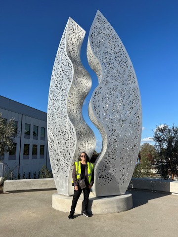Artist poses before a large-scale sculptural artwork with a bright blue sky