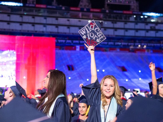 Woman in cap and gown at Commencement ceremony holding a cap that says Wildcat for Life