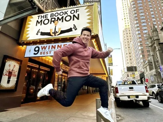 Photo of Alumni Tony Moreno ’21 jumping in front of a theater playing "The Book of Mormon" - the Broadway musical he is a "swing" on.