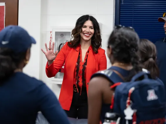 A photograph of a woman in a red blazer smiling and waving at student athletes 