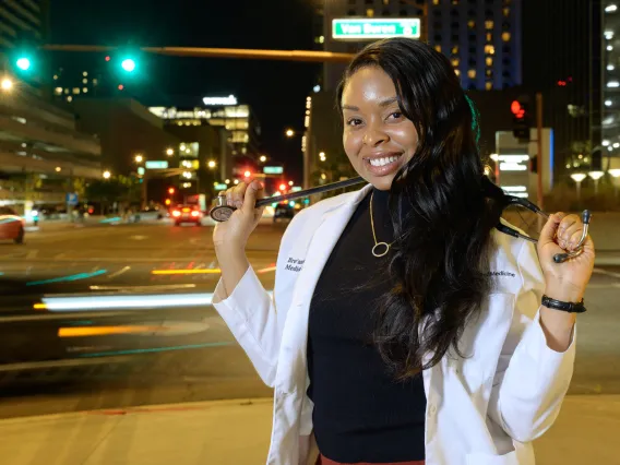 A photograph of a woman standing in a street wearing a white lab coat, holding a stethoscope around her neck. 