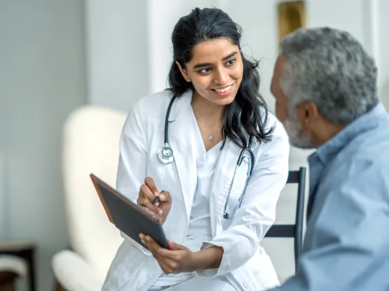 A photograph of a doctor wearing a white lab coat holding a tablet, showing it to an older adult patient. 