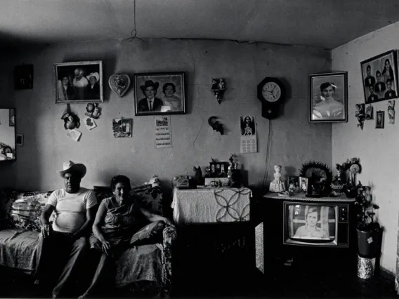 A photograph of a man and a woman sitting in a living room with family photos, a mirror, clock and television behind them.