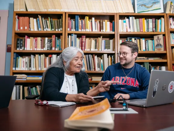 A woman talking and pointing to a computer while a man smiles at her in front of a shelf of books.