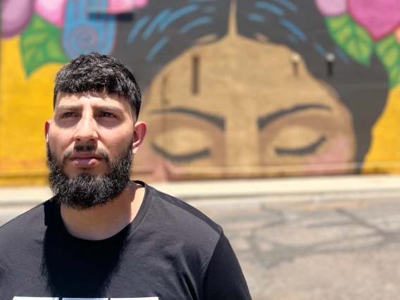 : A man stands in front of a colorful mural featuring closed eyes, flowers, and symbols on a sunny day.