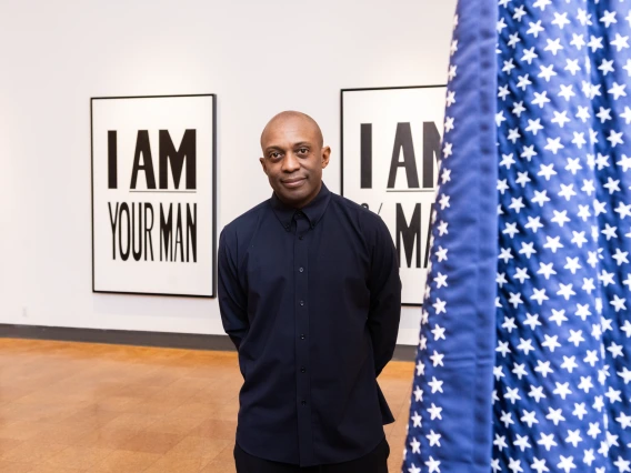 A man in dark clothing stands in a gallery between artworks that read “I AM YOUR MAN,” with a star-spangled fabric partially obscuring the frame.