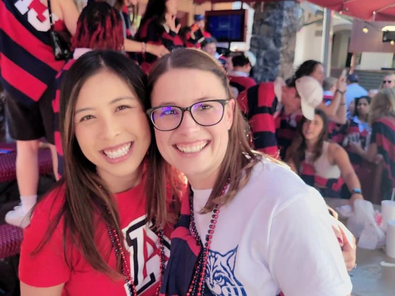 Two people wearing Arizona gear smile and pose together at a lively event filled with people in red and navy striped rugby shirts.