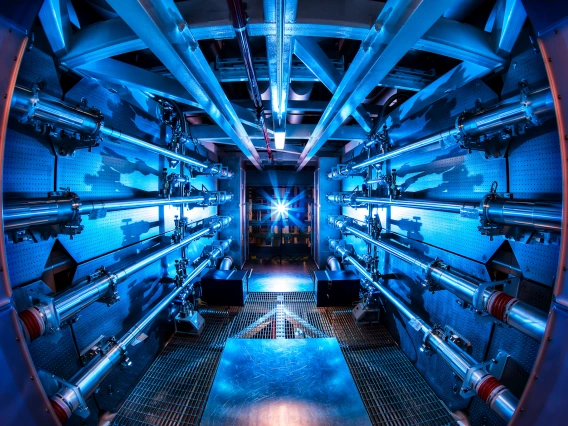 Interior view of a high-tech lab or facility glowing in blue light, showing symmetrical machinery with beams converging toward a central point.