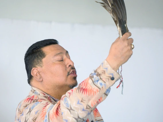 A person holding an eagle feather upright in a ceremonial gesture, wearing a patterned jacket and beaded necklaces, eyes closed in a moment of reflection.