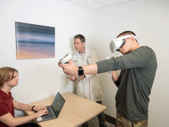 A man wearing a virtual reality headset holds VR controllers while a clinician in a lab coat observes and another person types on a laptop, in a clinical research setting.