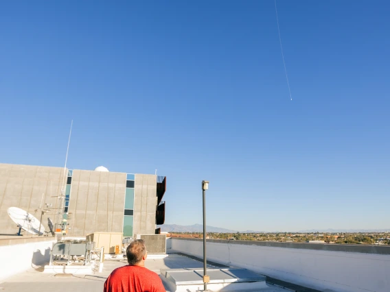A man in a red shirt watches a white weather balloon as it rises high into the bright blue sky above a rooftop filled with satellite dishes and equipment, with distant mountains in the background.