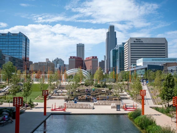 A panoramic view of Omaha’s city skyline with tall buildings, set against a mountain backdrop, with a calm lake in the foreground reflecting the soft hues of a cloudy sky.