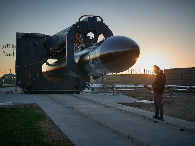 A person in a beanie stands with a laptop beside a black rocket secured in a large launch support structure, with the sun low on the horizon casting a warm glow across the industrial test site.