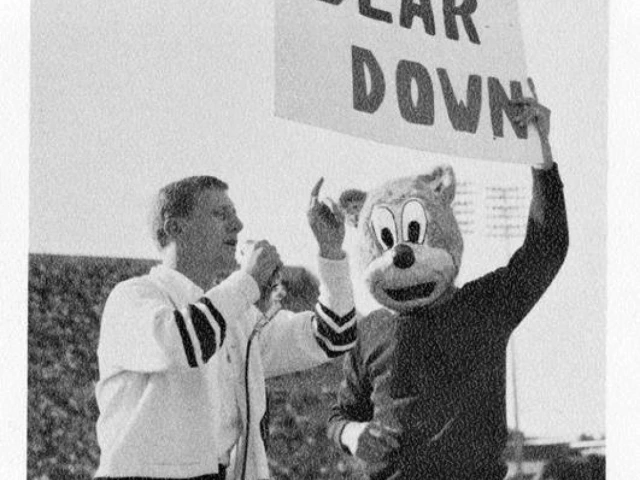 Historic black-and-white photograph of an early Wilbur Wildcat mascot standing beside a cheer leader holding a "BEAR DOWN" sign at a University of Arizona event.