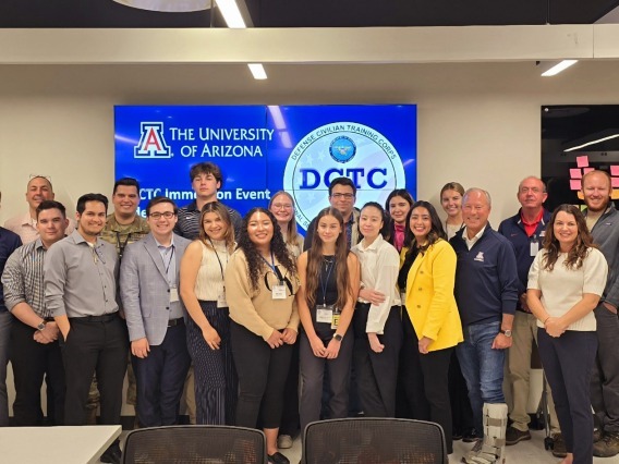 A group of students and teachers stand together for a group photo in front of a blue sign that says "The University of Arizona Defense Civilian Training Corps"