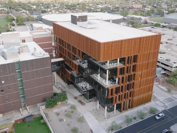 An aerial view of a copper-sided building with multiple levels of outdoor balconies. The building is attached to another via a walkway. Cars can be seen driving on the street outside.