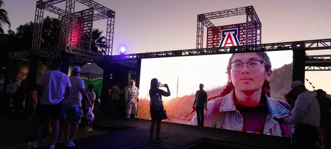 Event attendees watch large screen of video content on outdoor stage at sunset