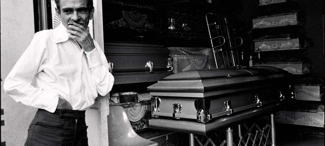 A man leaning against a wall with his hand on his mouth. A casket is in the background. 