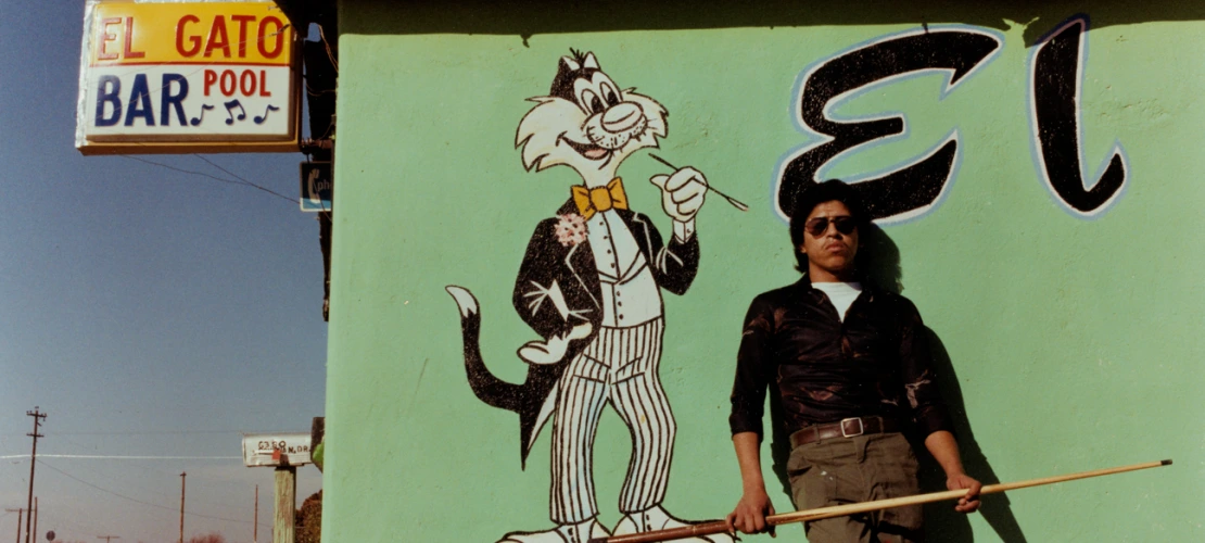 A photograph of a man standing with his back against "El Gato Bar and Pool" with a cue stick.