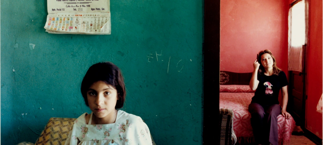 A photograph of a girl sewing with her mother in the room behind her smiling