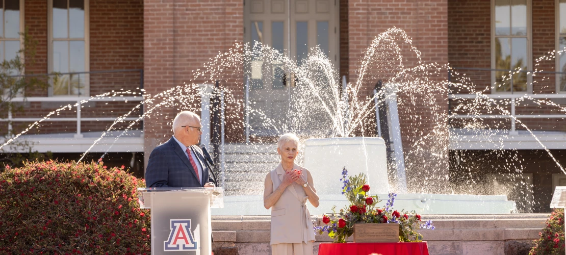 An image of Pat Gerleman standing in front of a crowd in front of the Old Main fountain. She has her arms over her heart in an expression of love and gratitude. Standing to her left, standing behind a clear podium with the University of Arizona block A logo on the front, is JP Roczniak, President and CEO of the University of Arizona foundation.