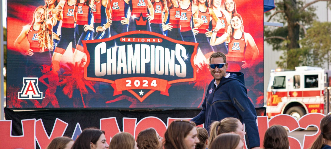 Coach and athletes celebrate in front of a large screen that reads “National Champions 2024” with the Arizona TriCats team.