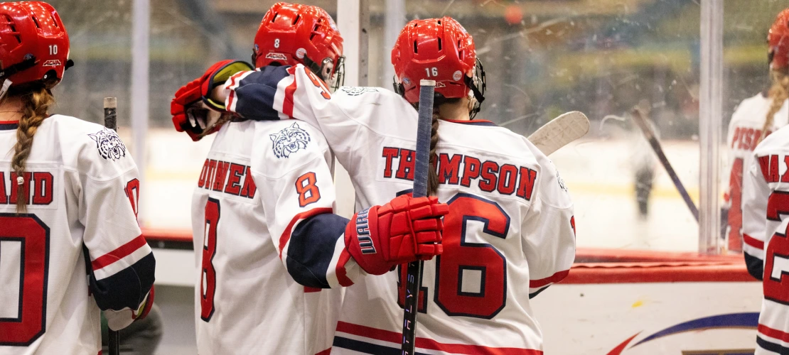 Two Arizona hockey players, with one labeled “THOMPSON,” stand arm-in-arm at the bench watching the rink.