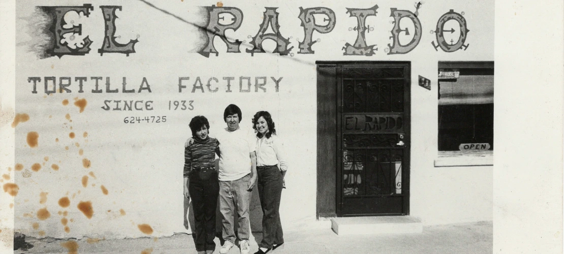 Three people standing and smiling with their arms around each other, outside a building that says "Tucson's Finest El Rapido Tortilla Factory Since 1933."
