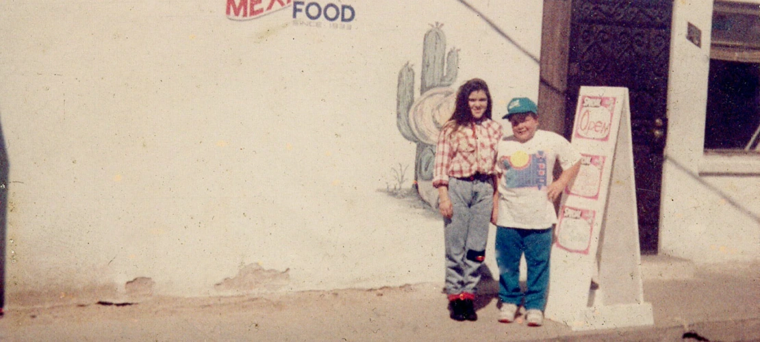 Two people standing and smiling outside of a restaurant that says "El Rapido Mexican Food."