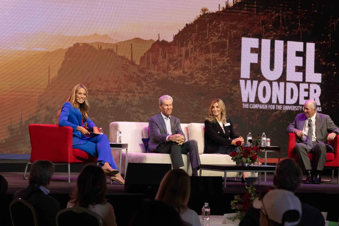 Woman in blue suit sits upon stage while 3 seated individuals talk.