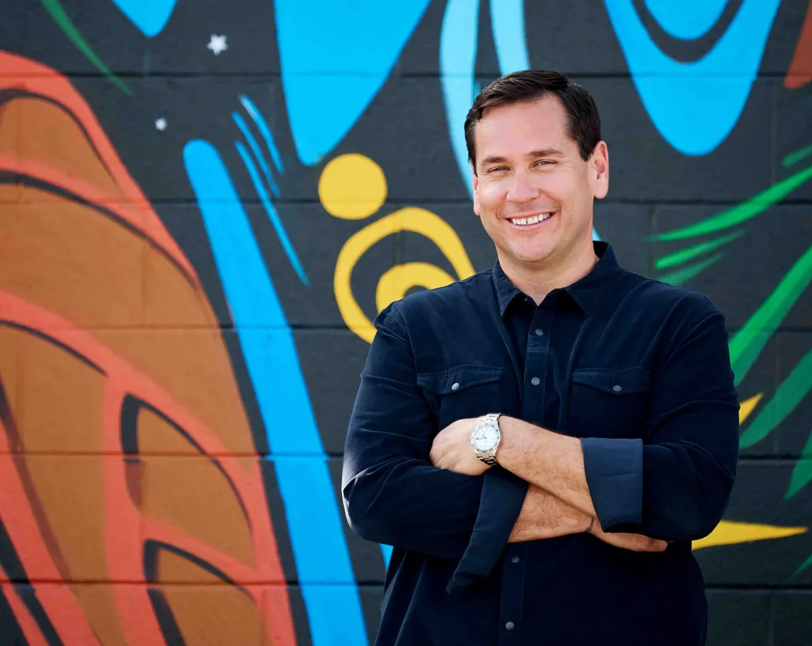 A smiling man crosses arms in front of brightly painted mural.