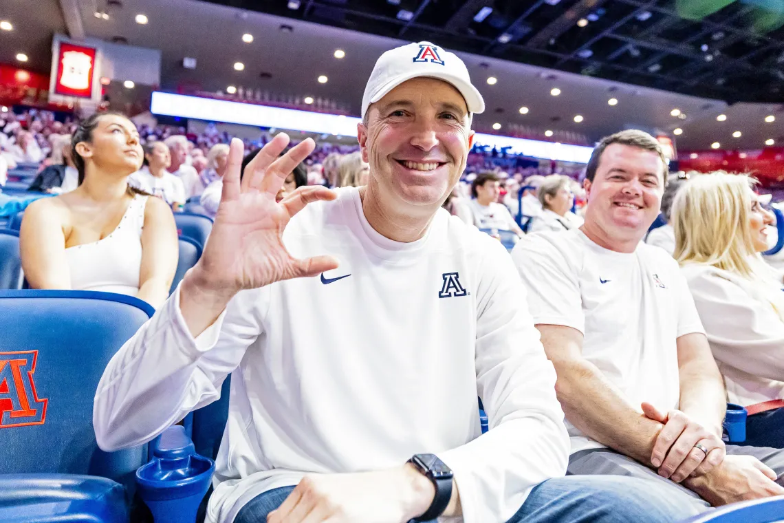 A photograph of a man wearing a white UArizona shirt and white UArizona hat holding up a Wildcat sign