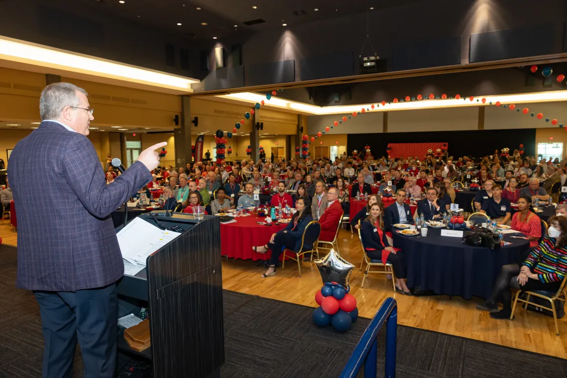 A presenter on stage talking to a crowd sitting on chairs around tables