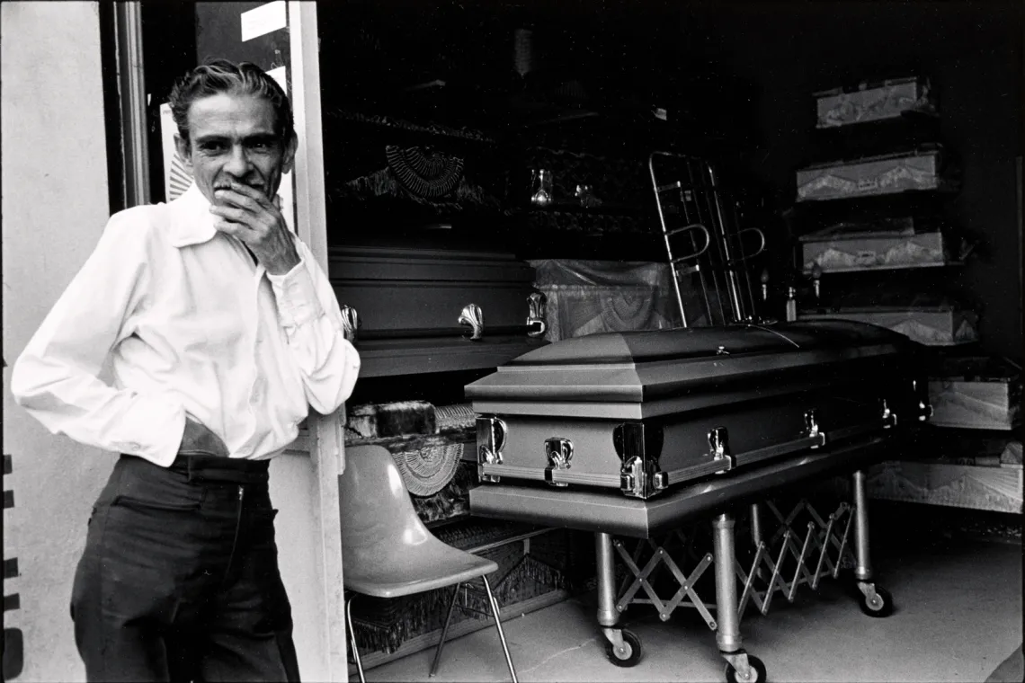 A man leaning against a wall with his hand on his mouth. A casket is in the background. 