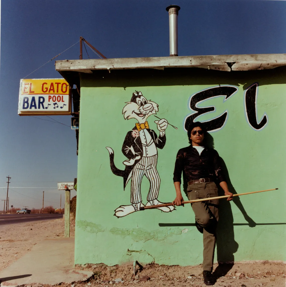 A photograph of a man standing with his back against "El Gato Bar and Pool" with a cue stick.