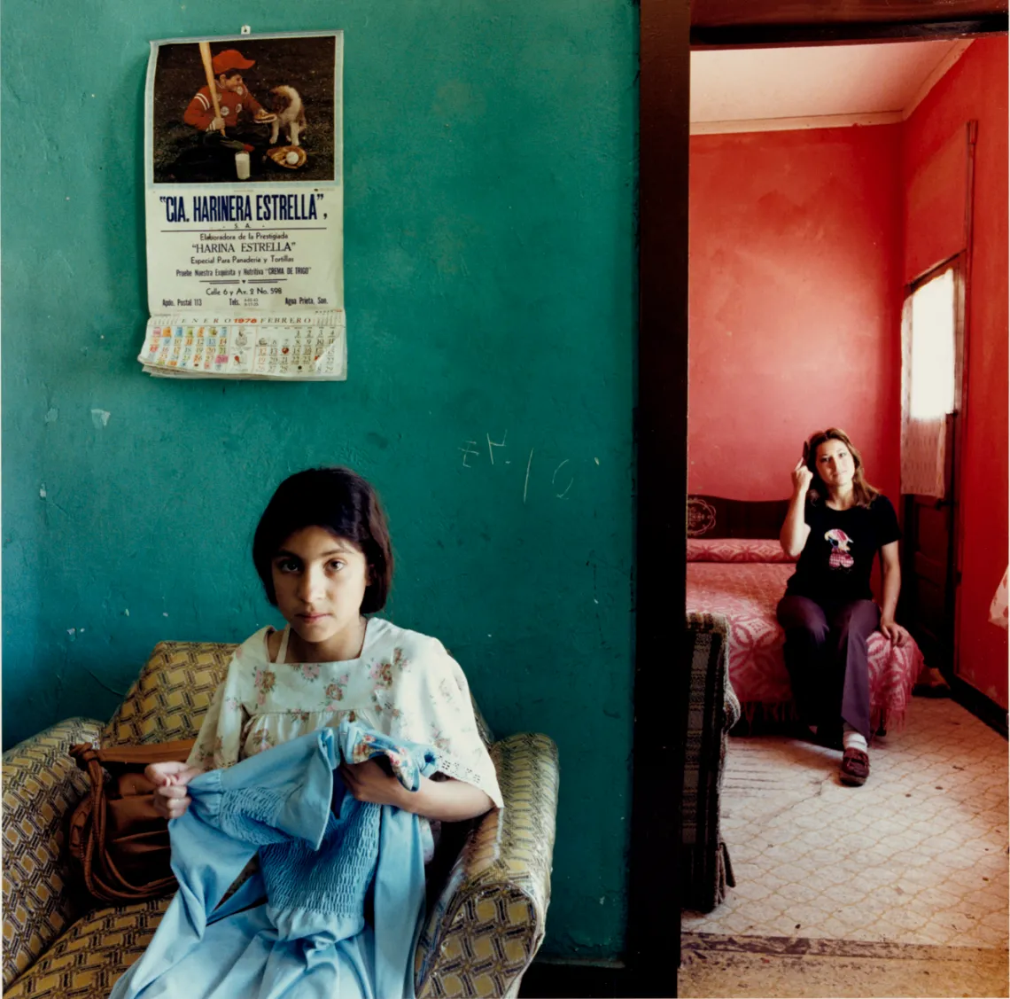 A photograph of a girl sewing with her mother in the room behind her smiling