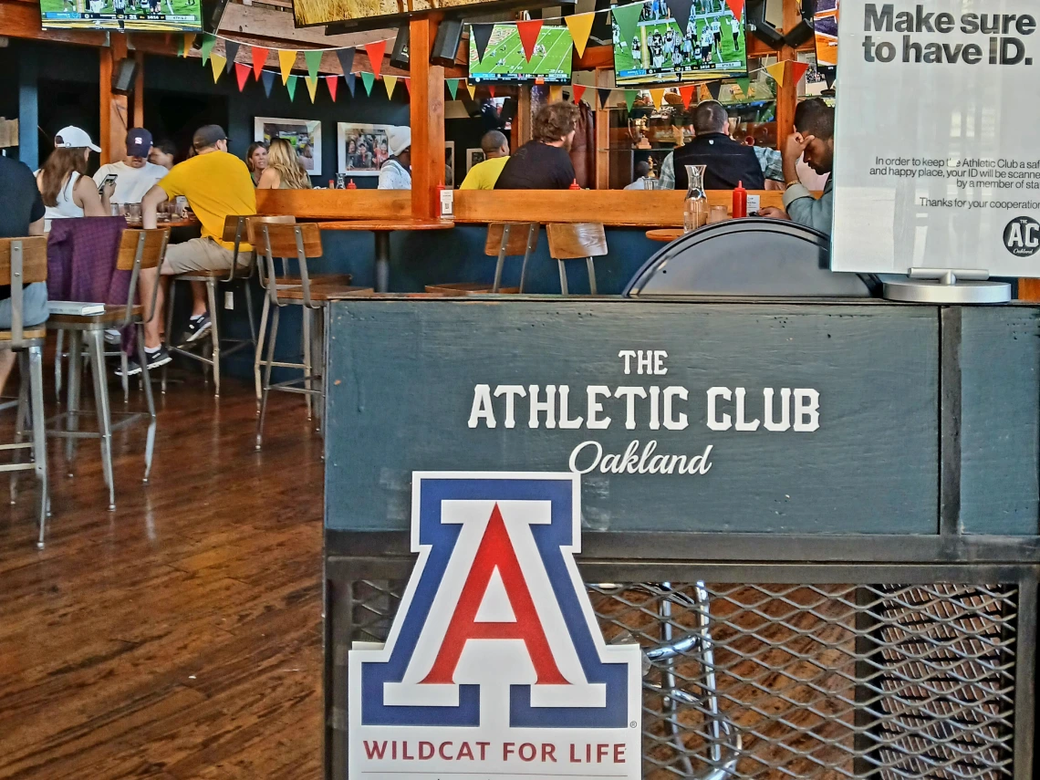 A bar with a University of Arizona Block A with people sitting and talking behind it, watching football