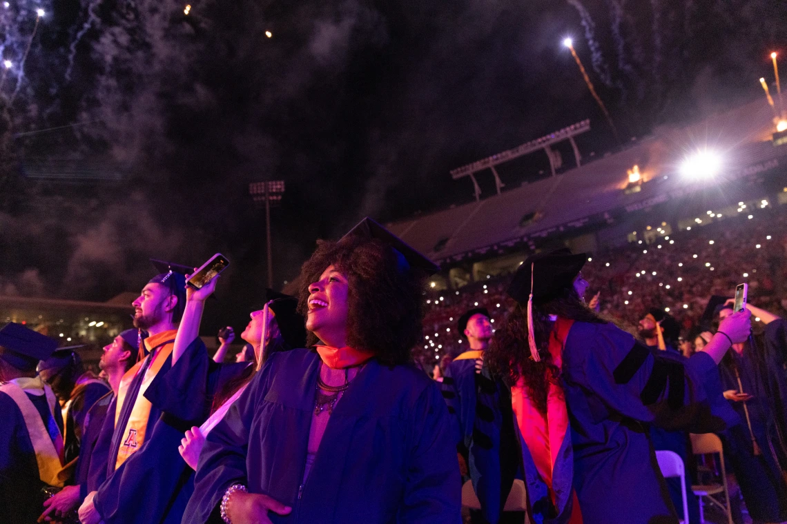 Graduates in academic robes look up at fireworks exploding above a stadium during a commencement ceremony.