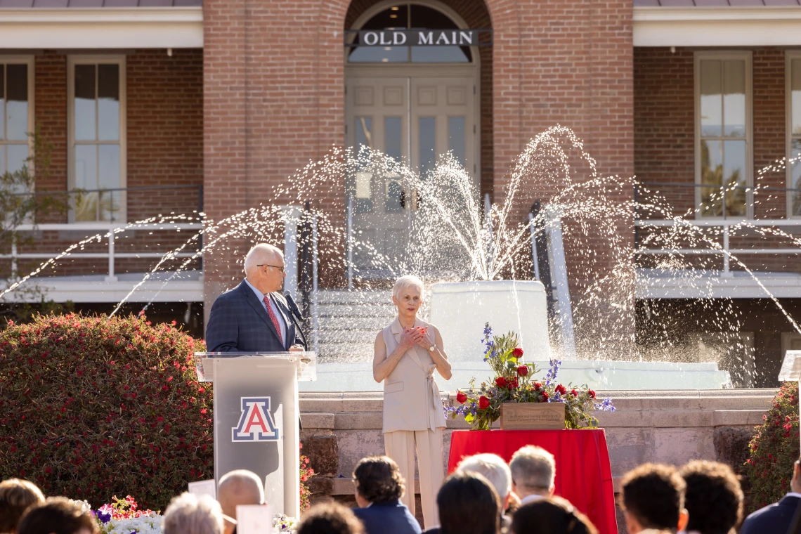 An image of Pat Gerleman standing in front of a crowd in front of the Old Main fountain. She has her arms over her heart in an expression of love and gratitude. Standing to her left, standing behind a clear podium with the University of Arizona block A logo on the front, is JP Roczniak, President and CEO of the University of Arizona foundation.
