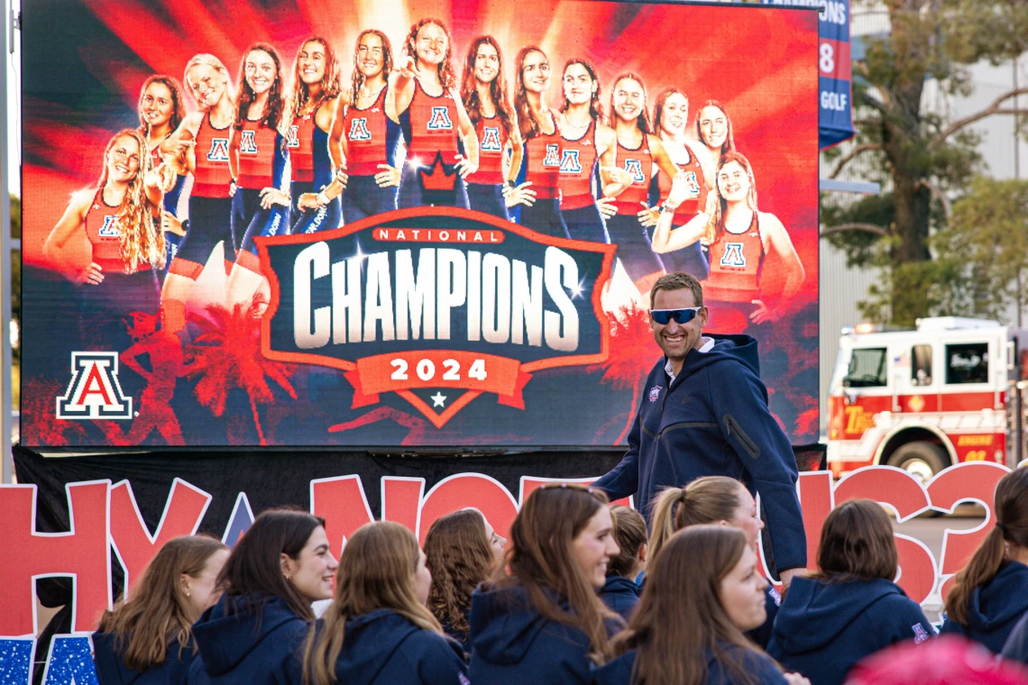 Coach and athletes celebrate in front of a large screen that reads “National Champions 2024” with the Arizona TriCats team.