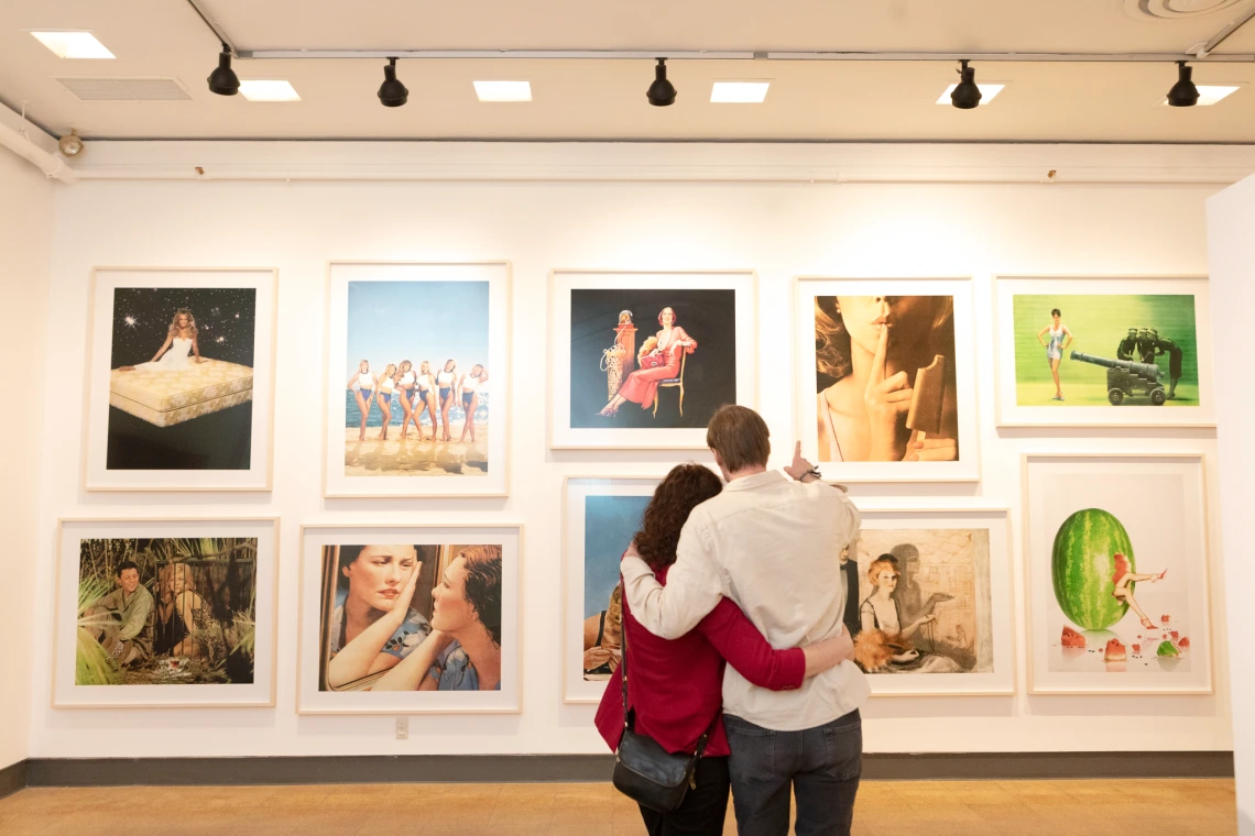 A couple stands closely, arm in arm, looking at a wall of colorful framed photographs in a well-lit art gallery.