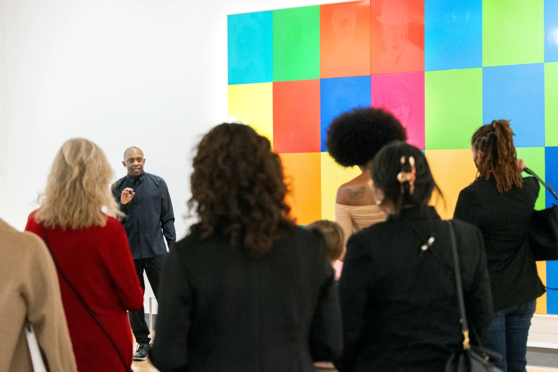 An artist speaks to a group of visitors in a gallery, standing before a wall filled with a grid of brightly colored, square portraits.