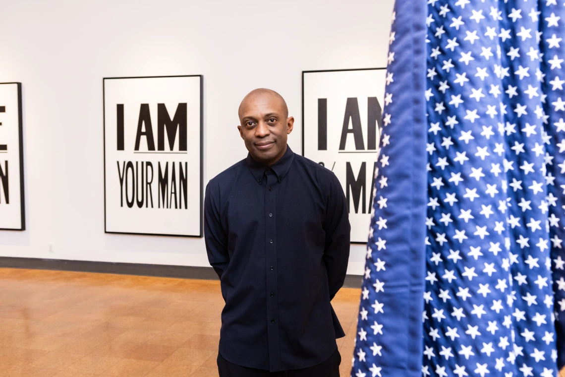 A man in dark clothing stands in a gallery between artworks that read “I AM YOUR MAN,” with a star-spangled fabric partially obscuring the frame.
