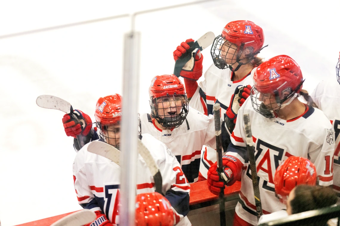 Arizona hockey players in red helmets gather on the bench, cheering and holding up their sticks.