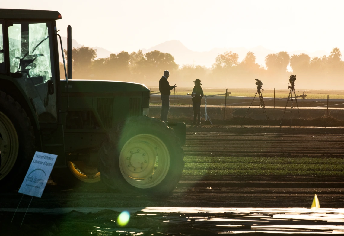 Silhouetted figures and camera equipment stand in a misty field at sunrise next to a green tractor, with trees and mountains in the background.