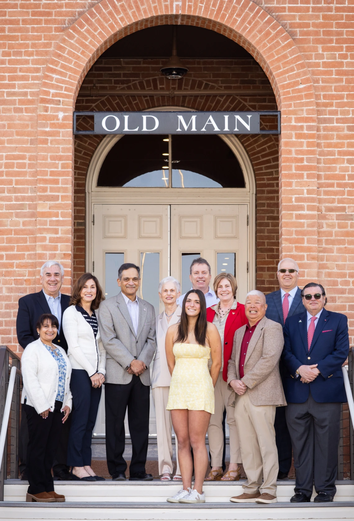 A group of twelve people pose smiling on the front steps of a brick building under a sign that reads “OLD MAIN,” with one young woman in a yellow dress standing at the front center.