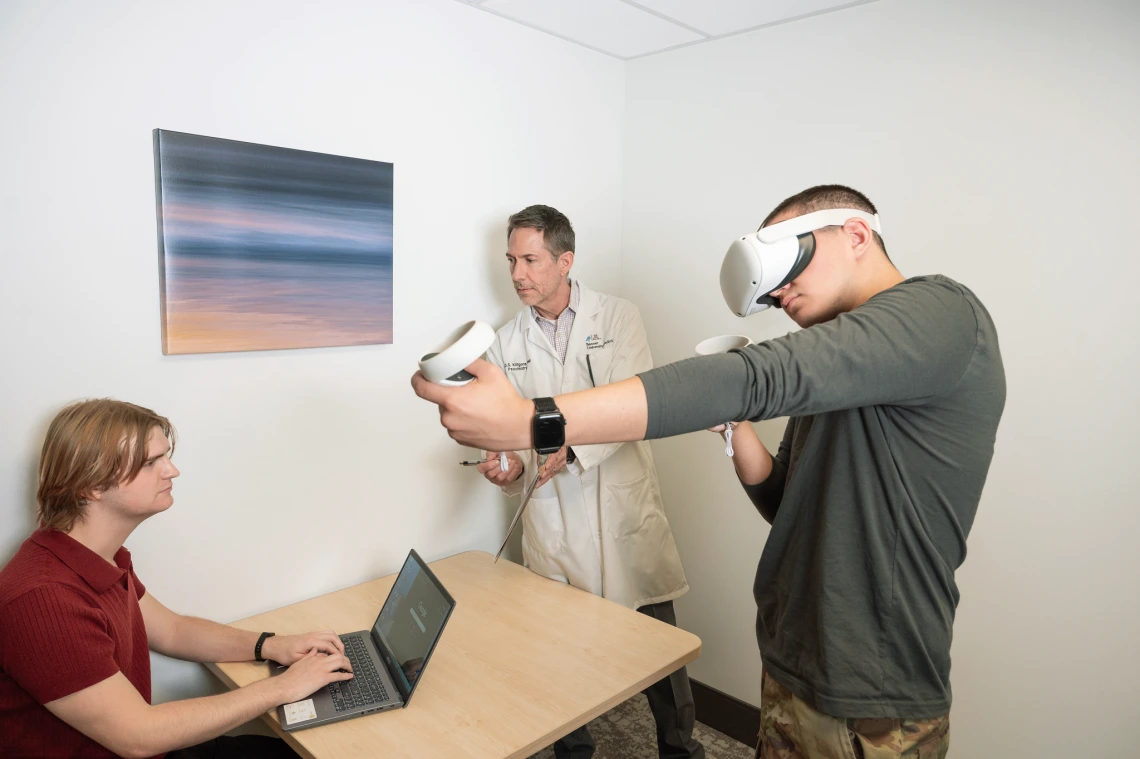 A man wearing a virtual reality headset holds VR controllers while a clinician in a lab coat observes and another person types on a laptop, in a clinical research setting.
