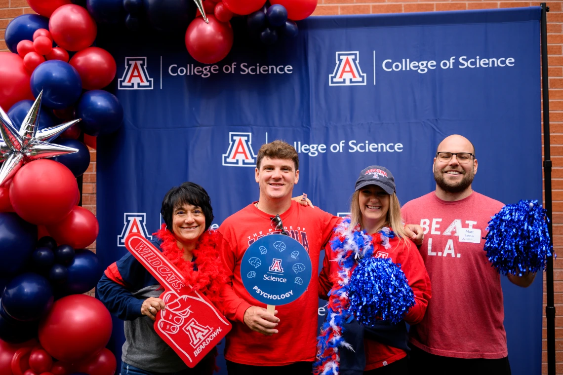 An image of four alumni (two women, two men) all smiling for a photo in front of the U of A College of Science backdrop. They are holding spirit items like a foam finger, a sign, and pom poms. Red and blue balloon trees are visible next to the group of alumni.