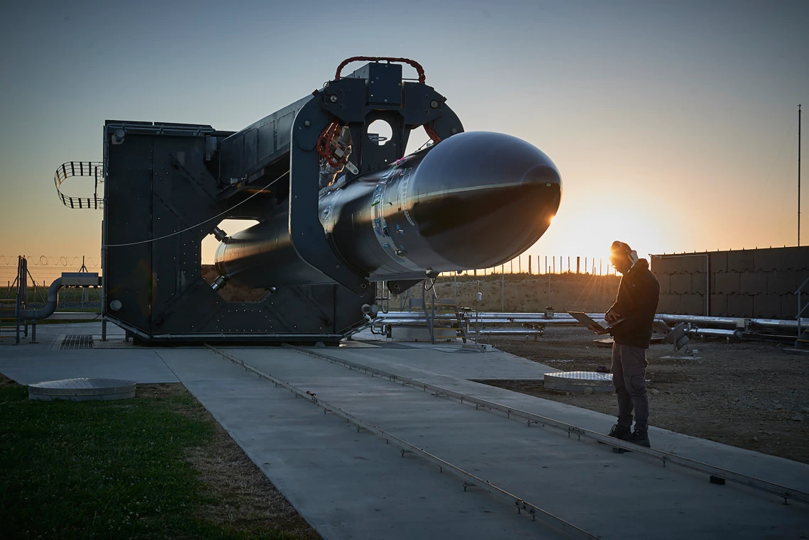 A person in a beanie stands with a laptop beside a black rocket secured in a large launch support structure, with the sun low on the horizon casting a warm glow across the industrial test site.