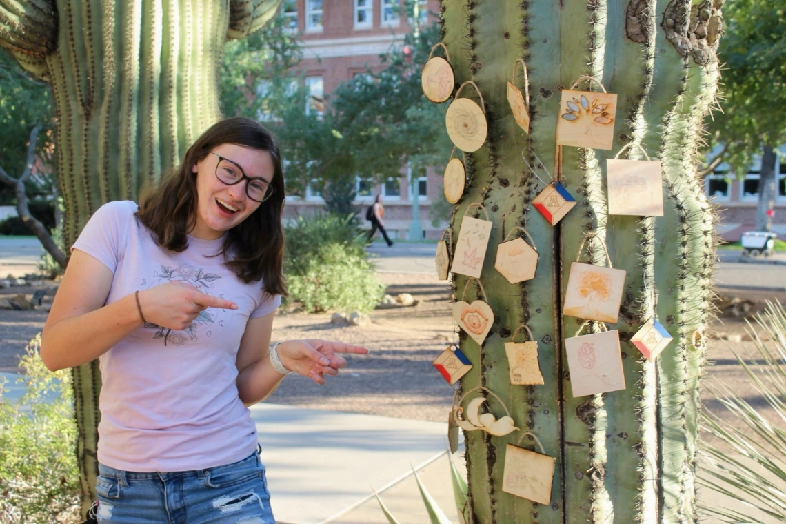 A girl standing and pointing to a cactus with lots of wooden art pieces.